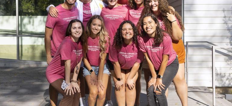 Students wearing Campus Ministry retreat shirts stand in two lines for a group photo outside of the Campus Ministry building.