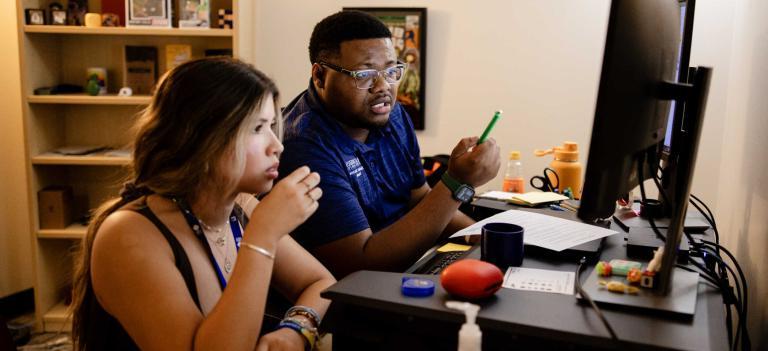 A success coach and a student meet in the success coach's office. They sit at the success coaches desk and look at the computer screen the success coach is pointing at with a pen.