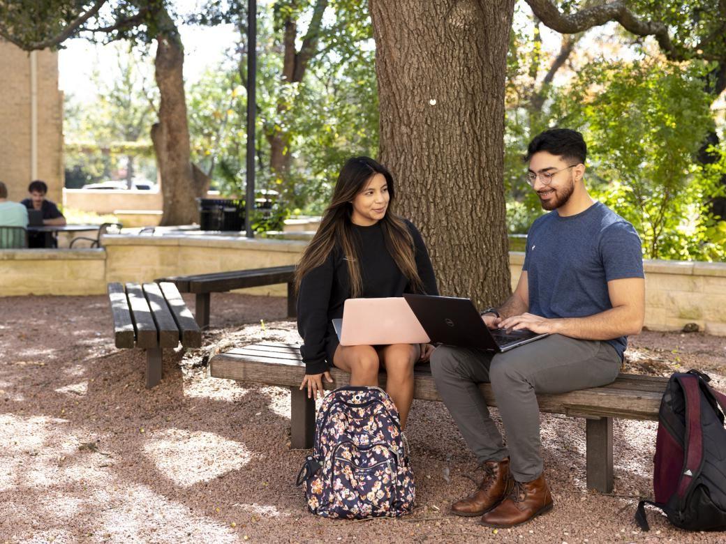 Two students sit under a tree on a bench on a patio with their backpacks on the ground next to them and work on their computers.