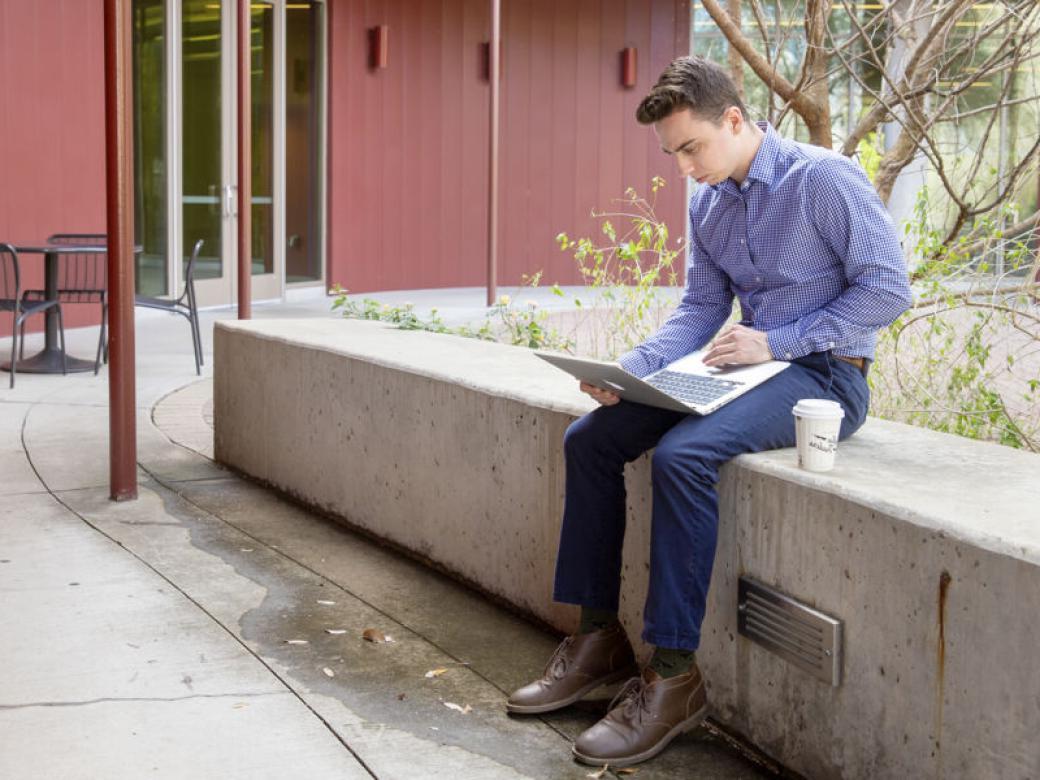 A graduate student sits on a concrete bench looking at their laptop with a cup of coffee next to them on the John Brooks Williams Natural Sciences Center - South patio.