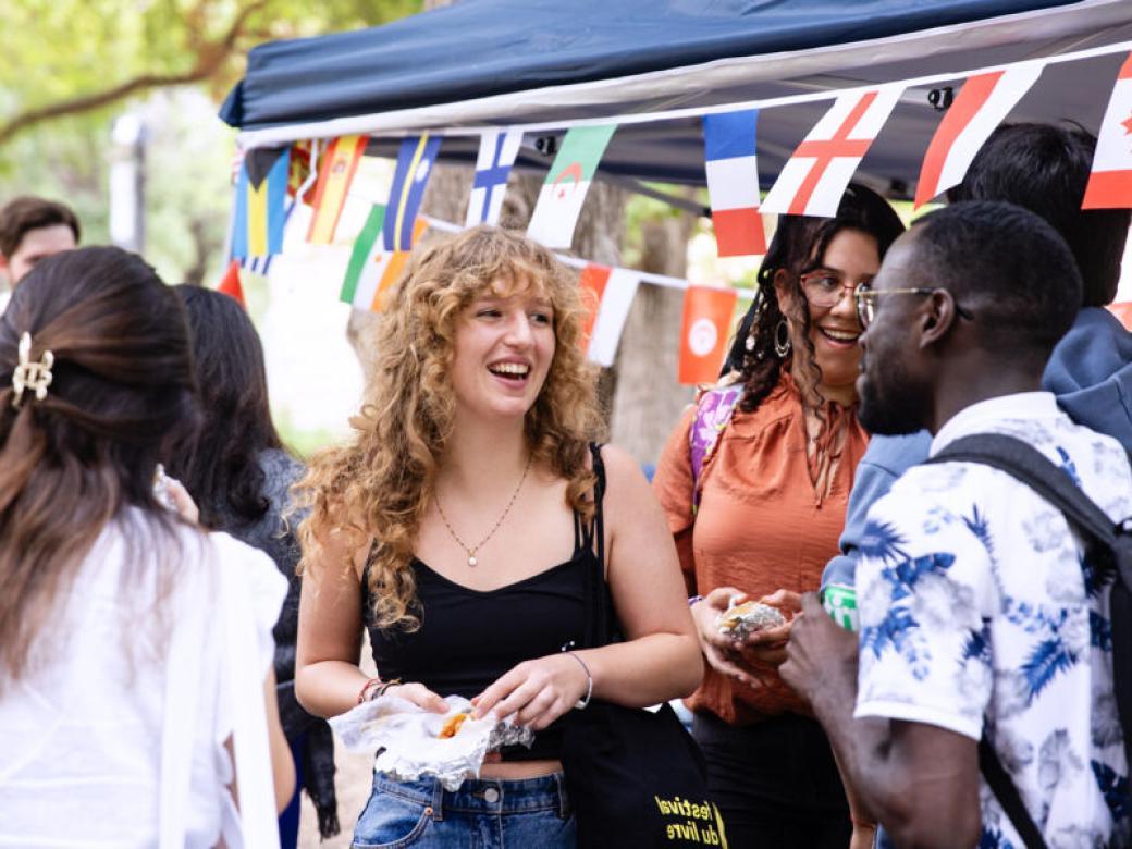 International students mingle and eat snacks by a tent displaying different countries' flags during an international student event.