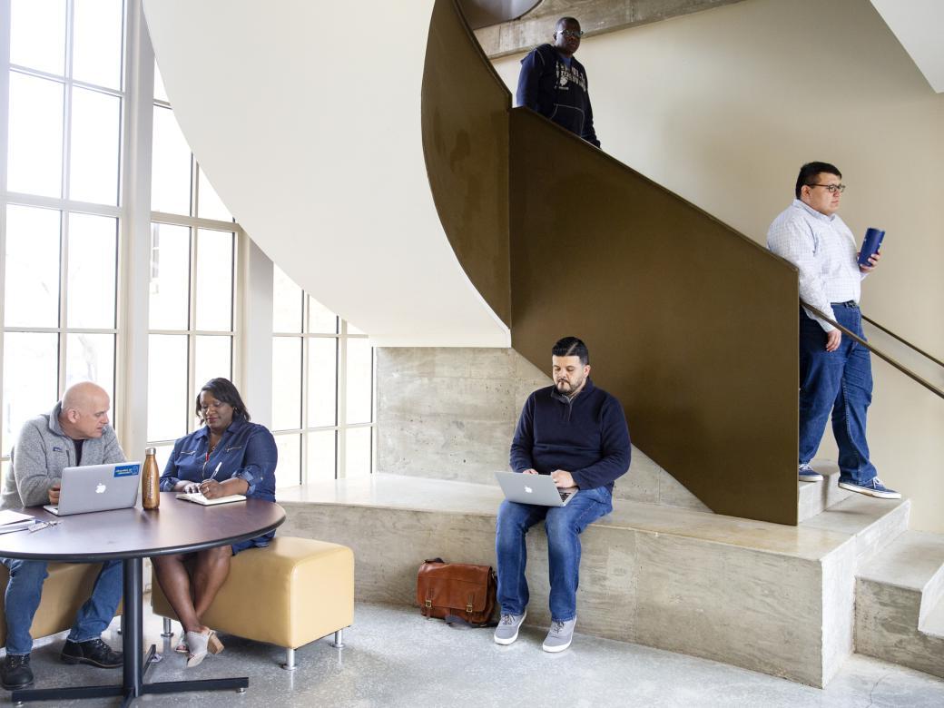 A student walks down the stairs. Another student sits on a bench. Two students sit at a table. All inside The Bill Munday School of Business.