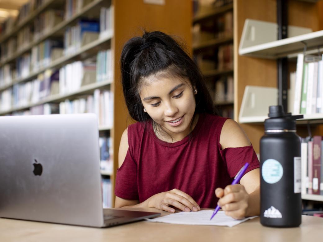 A student sits at a table with a water bottle and a laptop and takes notes on a piece of paper. Bookcases are in the background.