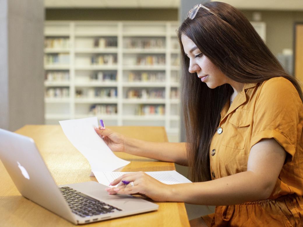 A student sits at a table holding a piece of paper and a pen as they look at their laptop. Bookcases are in the background.