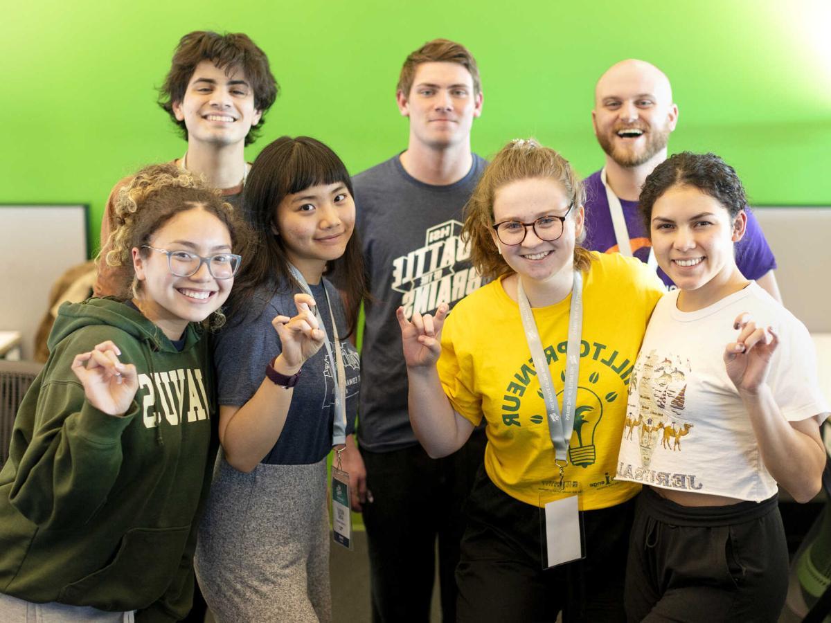 Seven students stand in front of a green wall and give toppers up hand signs.