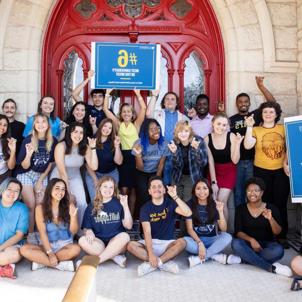 A group of St. Edward's University students pose in front of the red doors with the university president holding up signs celebrating the #6 best university in the west ranking