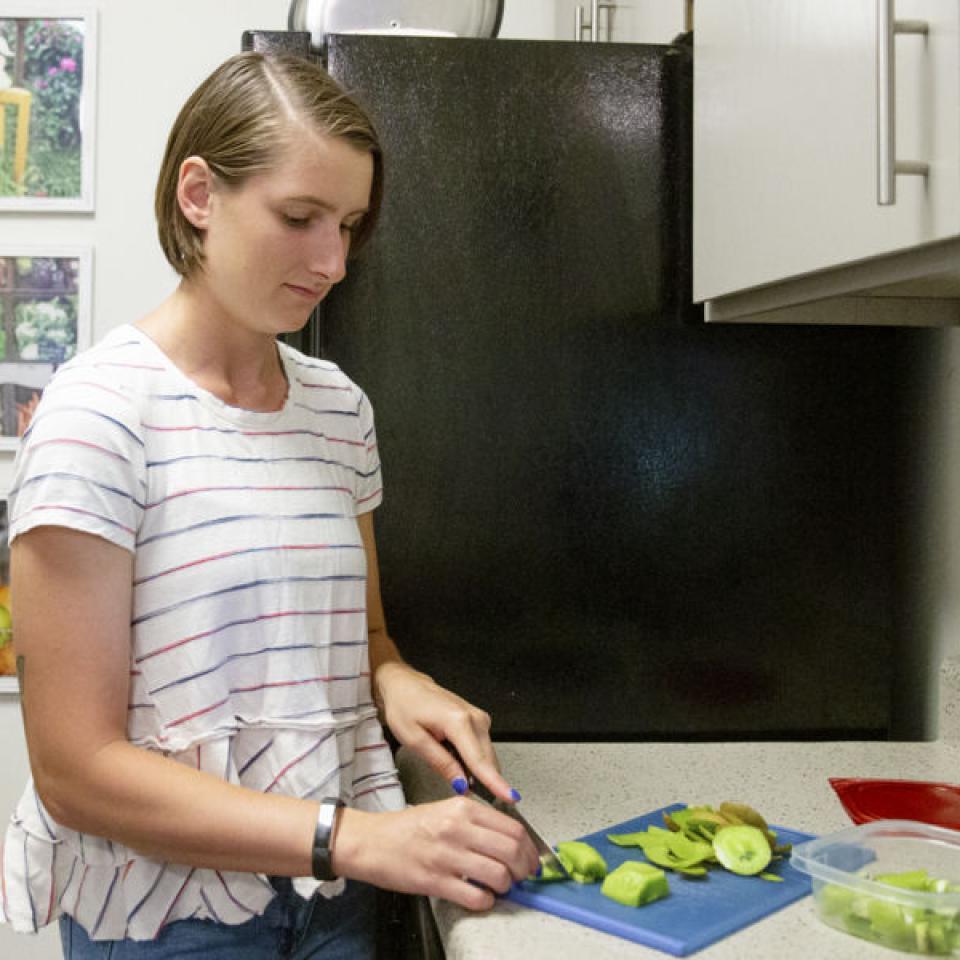 This image shows a woman with short brown hair, wearing a white blouse with red and blue stripes. 她正站在厨房里, slicing green vegetables on a blue cutting board placed on a countertop. A container with chopped vegetables and a red lid is nearby. 她身后是一个黑色的冰箱, and the wall has a series of colorful framed photographs depicting various fruits and vegetables. The setting appears to be a well-lit, clean, and organized kitchen.