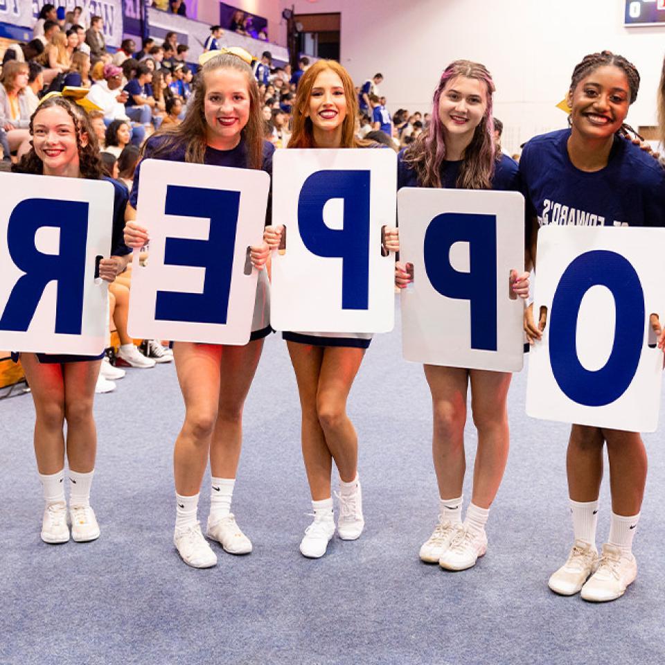 这张照片显示了一群七名妇女站在健身房, holding large signs that spell out "TOPPERS." 他们都在微笑 and wearing matching "St. Edward's Hilltoppers" T-shirts and athletic shoes. 这里的环境让人联想到赛前动员会或学校精神活动, 后面坐着一群人, 为活动欢呼和参与. 气氛充满活力和庆祝.