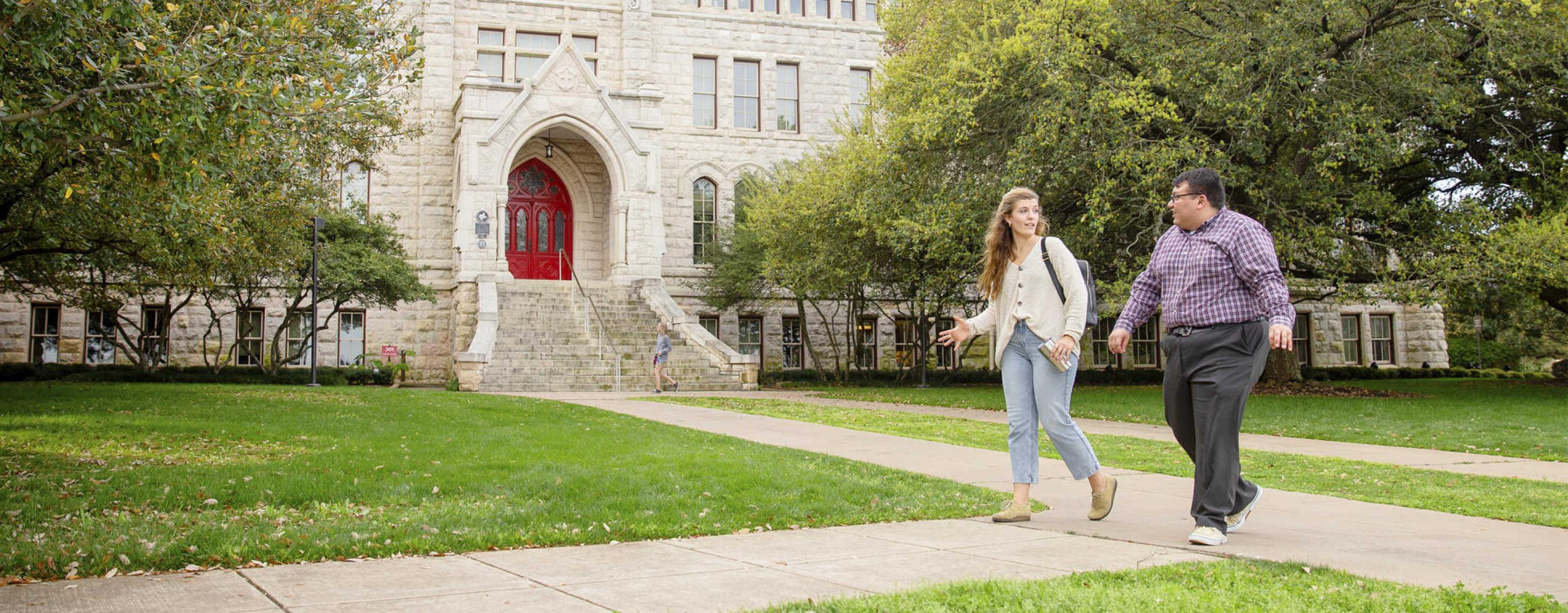 Two graduate students walk on a path in front of Main Building and oak trees.