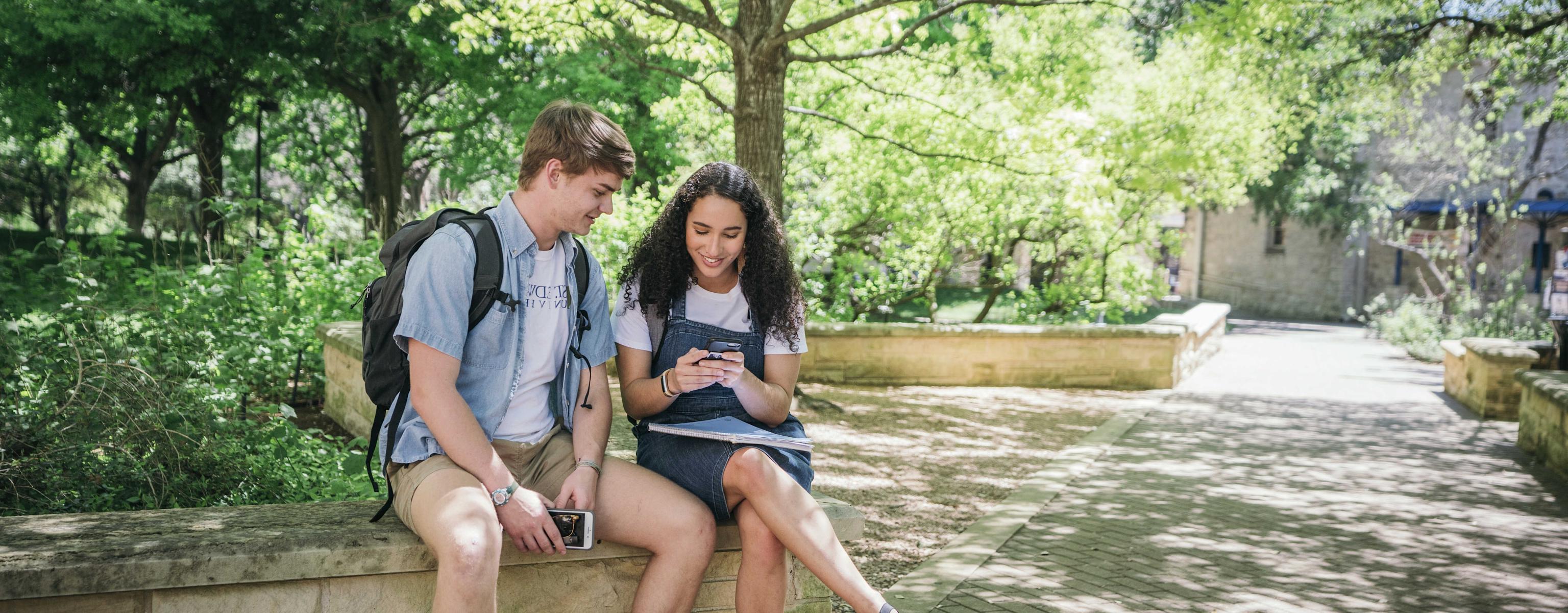 two students sit on a stone bench by a tree-lined walkway and look at a phone.