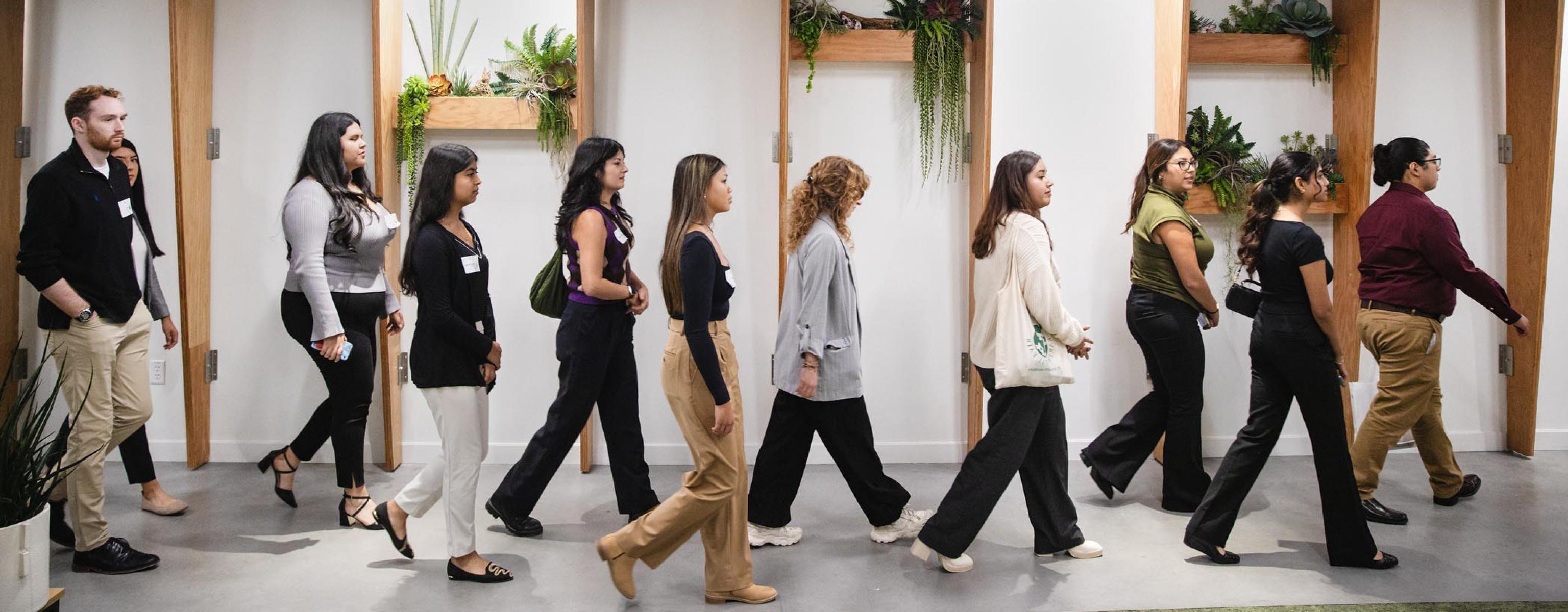 The image shows a group of professionally dressed individuals walking in a single file line indoors. The setting appears to be modern, with wooden beams and minimalistic design elements. The background features decorative shelves with plants, adding a touch of greenery to the space. The group consists of men and women, likely participating in a tour, workshop, or professional development event. The atmosphere is organized and focused.