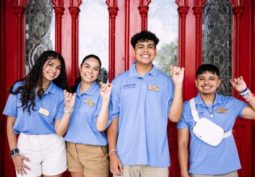 Four student orientation leaders stand in front of red doors wearing matching blue polos and make toppers up hand signs.