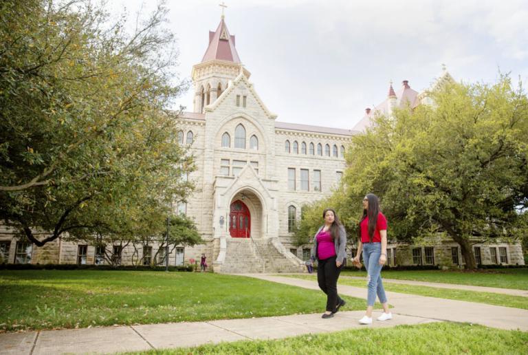 Two students walk on a path in front of Main Building and oak trees.