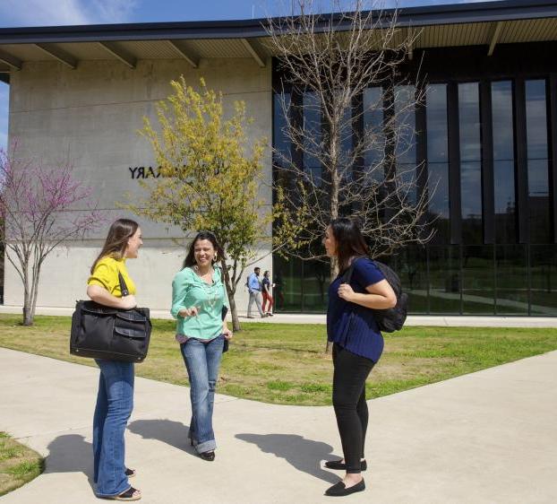 Three graduate students stand together on a pathway outside of Munday Library and chat.