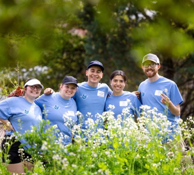 Five students wearing blue shirts stand together, framed by greenery, in the Students for Sustainability campus garden.