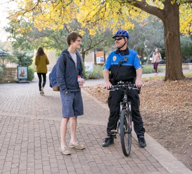 A university police officer sits on their bicycle and talks to a student as other students walk behind them.