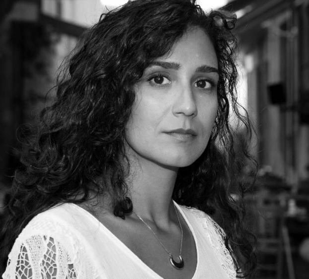 This is a black 和 white portrait of a woman with curly, 黑发, gazing directly at the camera with a serious expression. 她穿着一件浅色衬衫，袖子上有花边，戴着一条新月形吊坠的项链. The background appears to be an outdoor setting, slightly out of focus, with what might be trees 和 buildings in the distance. The overall mood is calm 和 introspective.