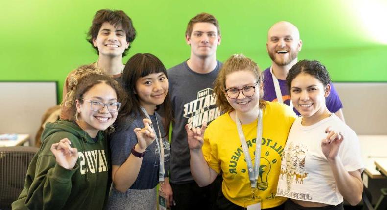 Seven students stand in front of a green wall and give toppers up hand signs.