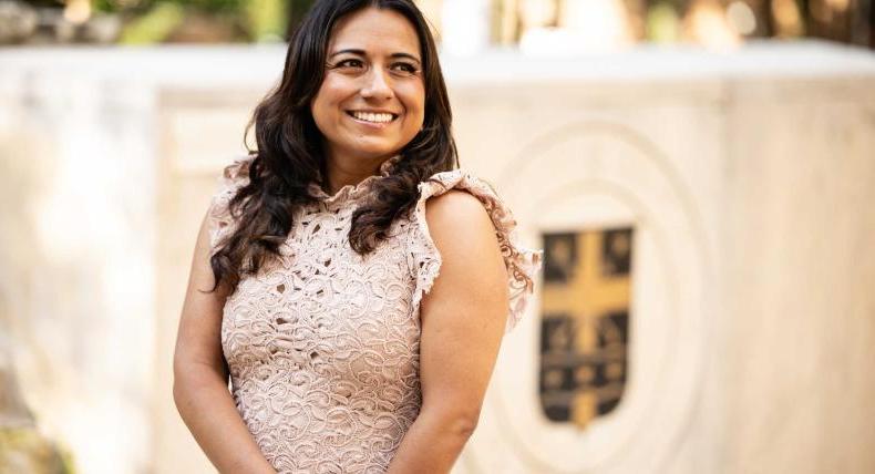 The image shows a woman with long dark hair wearing a light pink lace dress, smiling warmly while standing outdoors. In the background, there is a blurred emblem or crest on a stone structure. The setting appears to be a formal or professional environment, possibly a campus or an institution. The woman looks composed and confident.