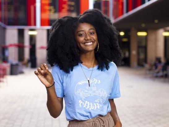 A student wearing a Welcome to the Hilltop shirt holds up a toppers up hand sign and stands in The Village patio area.