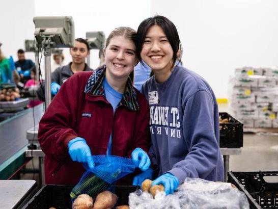 Two students hold produce and smile while volunteering at a food bank.