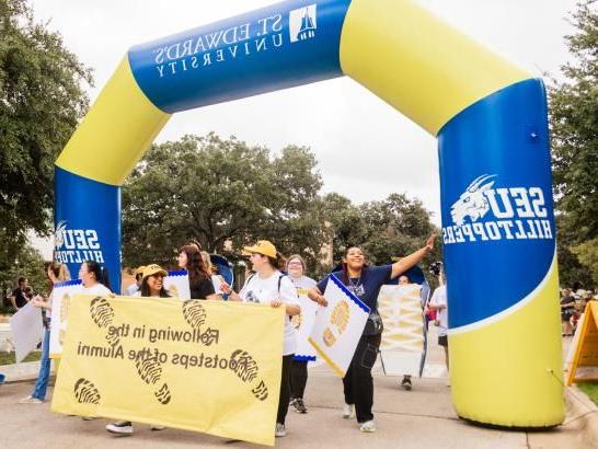 Following in the Footsteps of 校友 students walk with a banner displaying the same name under a blue and gold inflatable arch during the homecoming parade.