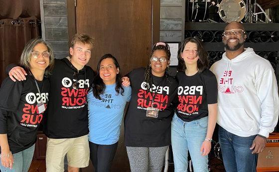 The image shows a group of six people standing together indoors, smiling at the camera. They are wearing casual clothing, including T-shirts that read "PBS NewsHour" and "Be The Change." The group includes both men and women, suggesting a diverse team. The background features musical instruments, indicating the setting might be a music room or studio. The atmosphere appears friendly and collaborative, likely related to a project or event focused on journalism or social impact.