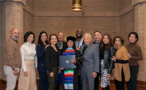 The image shows a group of twelve people standing together indoors, celebrating an academic achievement. The central figure is wearing a graduation cap and gown adorned with a colorful stole, holding a diploma. The surrounding individuals are dressed in semi-formal attire, smiling, and posing for the photo. The setting appears to be a hall or auditorium with brick walls, indicating a formal graduation ceremony. The atmosphere is joyful and supportive.