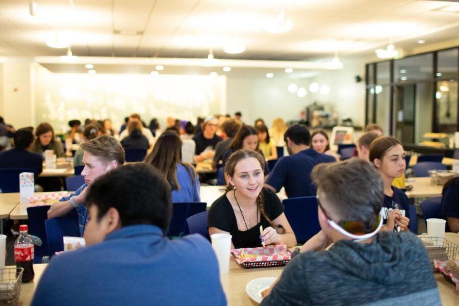 The image shows a bustling dining hall filled with college students. They are seated at tables, engaged in conversations while enjoying their meals. The atmosphere is lively and social, with groups of students interacting. The room is well-lit, featuring modern décor and a welcoming environment. 在前景, a young woman is smiling and talking with her peers, highlighting the communal and friendly vibe of the dining space.